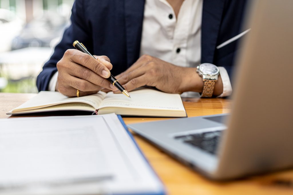 Stylized image of lawyer conducting research for a case. Close-up on hands writing in a journal in front of a computer